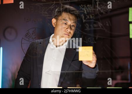 Scrum Kanban-Platine. Verschwommener Blick auf den nachdenklichen koreanischen Geschäftsmann in Anzug, der Geschäftsideen oder Aufgabenlisten auf Haftnotizen an der gläsernen Bürowand schreibt. Foc Stockfoto