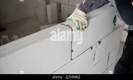 Ein Arbeiter baut aus einem Zellblock eine Wand. Nahaufnahme der Hände eines Baumeisters, die beim Bau einer Wand einen Block platzieren. Stockfoto