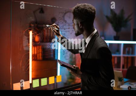Business and People Concept - afroamerikanischer Geschäftsmann, der im Büro auf Glastafel schreibt Stockfoto