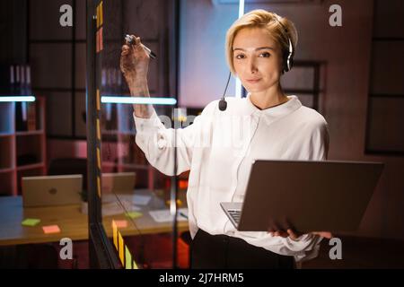 Seitenansicht einer fokussierten Arbeitnehmerin, die ein Headset trägt und einen Laptop verwendet, Ideen und Projektaufgaben an der Glaswand im dunklen Büro schreibt. Überarbeitungsverfahren bei Bürokonzept Stockfoto