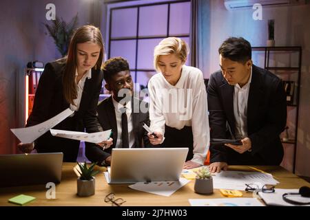 Geschäftsverhandlungen. Gruppe von verschiedenen modernen Menschen in formeller Kleidung mit Laptop und Finanzberichte bei der Sitzung. Kollegen, die am Schreibtisch stehen und im Vorstandszimmer Brainstorming durchführen Stockfoto