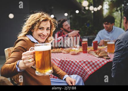 Porträt einer wunderschönen vierzig Frau, die die Kamera anschaut und Glas mit Lagerbier hält, während sie am Bauerntisch sitzt. Im Hintergrund eatin Freunde Stockfoto