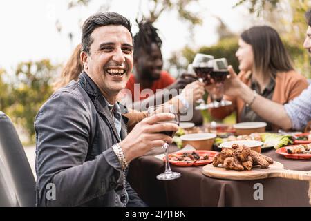 Porträt eines lächelnden, reifen Mannes, der die Kamera mit einem Glas Rotwein betrachtet - im Hintergrund verschwommen, toben Freunde der Familie zusammen - Stockfoto