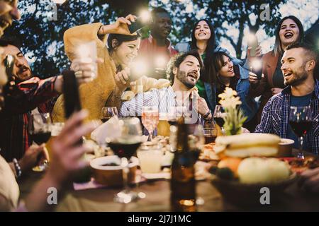 Gruppe betrunkener junger Menschen, die laut am Grilltisch singen, Wein und Bier trinken und Lichter mit Smartphones Fackeln machen - Freunde Stockfoto