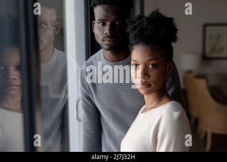 Porträt einer ernsthaften afroamerikanischen jungen Freundin mit ihrem Freund, der zu Hause am Fenster steht Stockfoto
