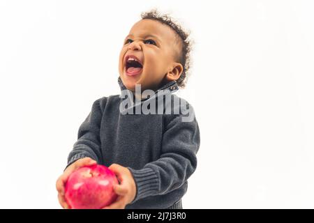 Netter multirassischer Junge, der sich weigert, seinen roten saftigen Apfel zu essen und über weißem Hintergrund schreit. Hochwertige Fotos Stockfoto