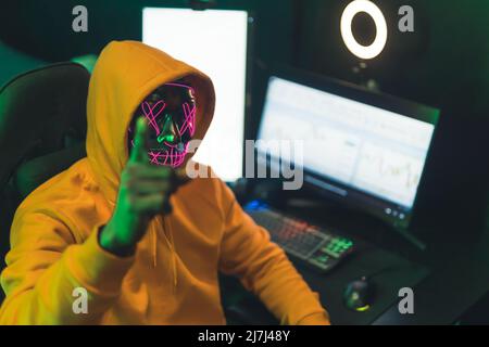 Mann mit einer gruseligen Neonmaske sitzt vor einem Computer-Hacking-Konzept-Medium aus der Nähe. Hochwertige Fotos Stockfoto