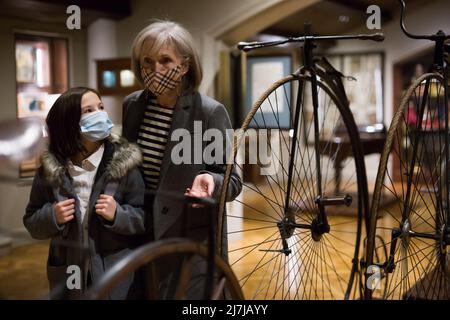 Zwischen einem Schulmädchen und einer älteren Tutorin in Gesichtsmasken beim Betrachten eines Vintage-Fahrrads im Museum Stockfoto