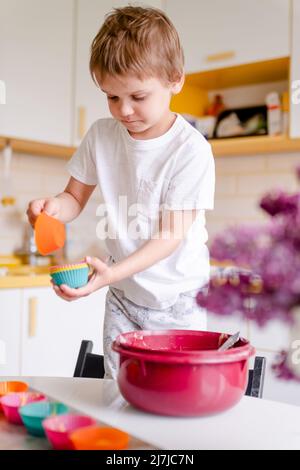 Kleines Kind 5 Jahre alt kocht Cupcakes in heller Küche zu Hause. Kleiner Junge verwendet Silikon-Backformen Stockfoto