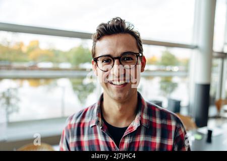 Porträt eines jungen Geschäftsmannes, der im Büro steht. Männliche Geschäftsexpertin in legerer Kleidung, die Brillen trägt und die Kamera anschaut und lächelt. Stockfoto