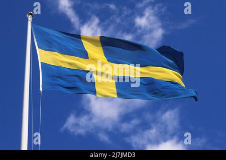 Nahaufnahme einer hochgesteppten schwedischen Nationalflagge, die im Wind gegen einen blauen Himmel mit hellen Wolken fliegt. Stockfoto