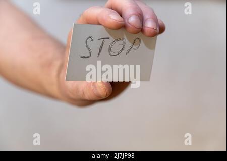 Stopp. Ein Wort, das in zackigen Buchstaben geschrieben wurde. Die Hand eines Mannes hält ein weißes Papierrechteck mit Text vor einem grauen Hintergrund. Stockfoto