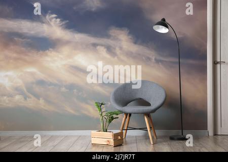 Stilvoller Sessel mit Zimmerpflanze und Lampe an der Wand mit bedrucktem Himmel im Zimmer Stockfoto