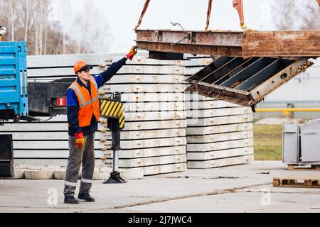 Slinger in Helm und Weste steuert das Entladen von Metallkonstruktionen auf der Baustelle. Weißer Handwerker entlädt Ladung. Stockfoto