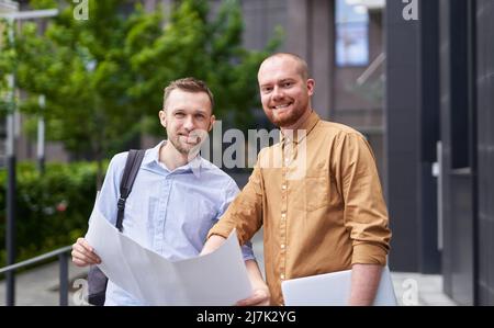 Selbstbewusstes Team aus Architekten oder Designer-Managern, das Zeichnungen oder Entwürfe hält, um das Projekt zu besprechen, Brainstorming zu veranstalten. Geschäftsteam im Freien in der Nähe des modernen Gebäudehintergrundes. Hochwertige Bilder Stockfoto