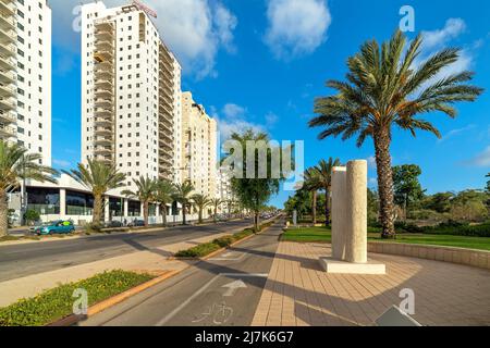 Bürgersteig und Stadtstraße entlang mehrstöckiger Wohngebäude unter blauem Himmel in Ashkelon, Israel. Stockfoto