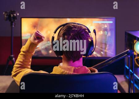 Rückansicht des professionellen Spielers, der auf einem Spielstuhl sitzt und seinen Sieg beim Online-Esport-Turnier feiert, macht eine Geste der Gewinnerhand, trägt ein Headset und schaut auf den Monitor. Neonlicht. Cyber-Sport Stockfoto