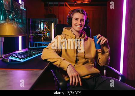Porträt eines lächelnden Pro-Gamer, der ein Headset trägt, die Kamera anschaut, das Energie-Koffein dink hält, auf einem Spielstuhl in der Nähe eines professionellen Gaming-Setups sitzt. Cyber-Sportler spielen Shooter Computerspiel Stockfoto