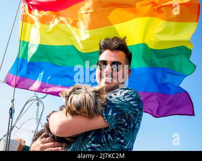 Gutaussehende schwule Person umarmt seinen kleinen Hund yorkshire Terrier auf einer Regenbogenfahne auf dem Hintergrund Stockfoto