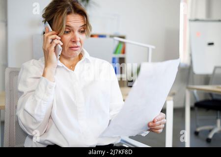 Empörte Büroangestellte, die Dokumente ansah und auf dem Smartphone sprach Stockfoto