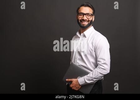Erfolgreicher indischer Geschäftsmann in formeller Kleidung und Brille mit Laptop-Computer, isoliert auf schwarzem Hintergrund stehend, Porträt des stolzen hispanischen männlichen Unternehmers Stockfoto
