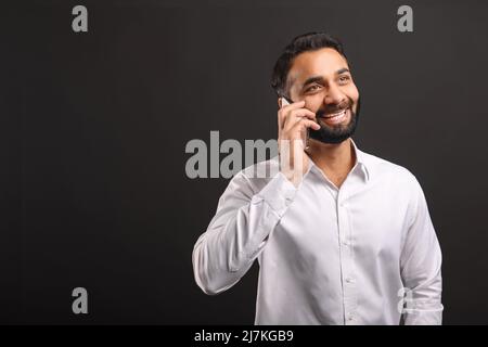 Glücklicher bärtiger indischer Mann im formellen weißen Hemd, der auf dem Smartphone auf schwarzem Hintergrund isoliert spricht. Männliche Büroangestellte genießen angenehme Telefongespräche, chatten auf dem Handy Stockfoto