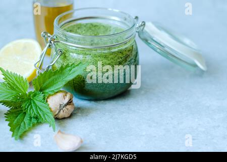 Vegetarische Brennnessel, Knoblauch, Olivenöl, Zitrone und Walnuss. Offenes Glas mit hausgemachtem Brennnesselpesto auf grauem Hintergrund. Veganes Brennnesselpesto Stockfoto