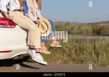 Die Beine zweier nicht erkennbarer Freundinnen mit einem Hut in den Händen sitzen im Lastwagen des weißen Autos. Bewundern Sie den Sonnenuntergang in einer ländlichen Landschaft Stockfoto