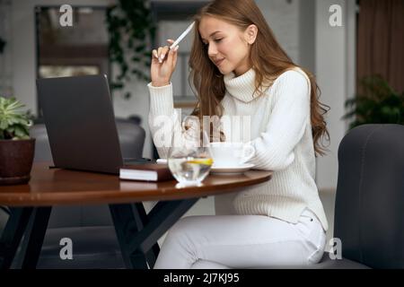 Schöne Frau, die am Tisch sitzt und mit einem Laptop ein Buch liest und im Internet surft Stockfoto