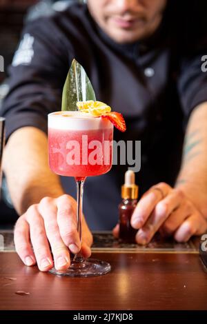Der männliche Barkeeper blickt auf die Kamera und serviert während der Arbeit in der Bar einen köstlichen exotischen schaumig-schäumenden Cocktail auf der Holztheke Stockfoto