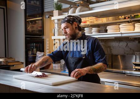 Fokussierter tätowierte Chefkoch in Schürze, der rote Zwiebeln mit Messer hackt, während er in einer hellen Küche kocht Stockfoto