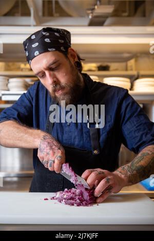 Fokussierter tätowierte Chefkoch in Schürze, der rote Zwiebeln mit Messer hackt, während er in einer hellen Küche kocht Stockfoto