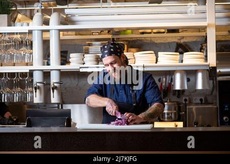 Fokussierter tätowierte Chefkoch in Schürze, der rote Zwiebeln mit Messer hackt, während er in einer hellen Küche kocht Stockfoto