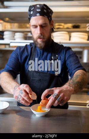 Fokussierter bärtiger männlicher Koch in Uniform, der rohes Ei in eine Schüssel zerbrach, während er am Tisch in der hellen Küche des Restaurants arbeitete Stockfoto