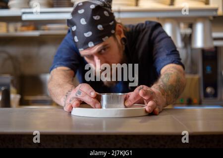 Fokussierter bärtiger Chefkoch mit Tattoos, der Metallringform auf weißem Teller anordnete, während er in der hellen Küche am Tisch stand Stockfoto