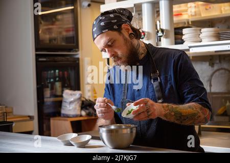 Fokussierter tätowierte männlicher Koch in Uniform, der frische Zutaten in die Schüssel gab, während er am Tisch in der hellen Küche des Restaurants arbeitete Stockfoto