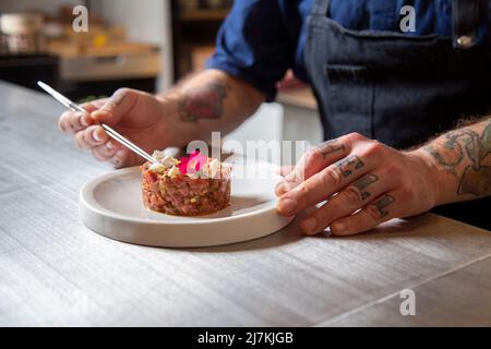 Zugeschnittenes, unkenntlich fokussiertes Männchen auf der Schürze, das frische, schmackhafte Rindertartare auf dem Teller in der Küche des Restaurants schmückt Stockfoto