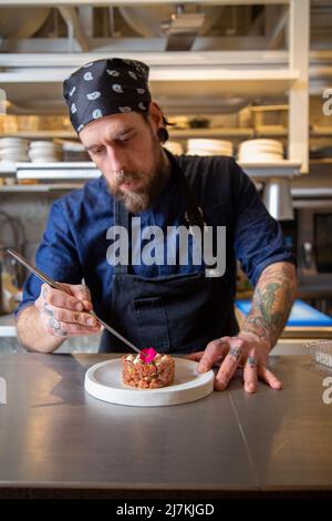 Gebärtete Chefköchin auf der Schürze, die frischen, leckeren Rindertartar auf dem Teller in der Küche des Restaurants verschönert Stockfoto