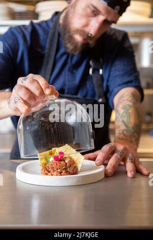 Der auf Getreide ausgerichtete männliche Koch in Uniform überdeckt während der Arbeit im Restaurant frische, leckere Tartare mit Glaskuppel am Tisch Stockfoto