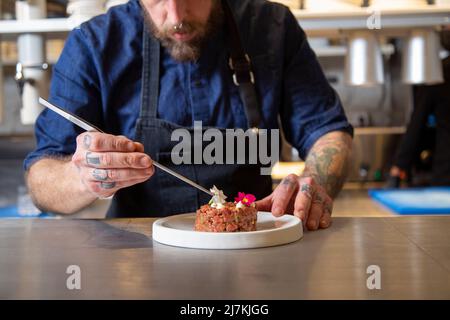 Cropped unkenntlich fokussierten bärtigen männlichen Koch in Schürze Dekoration frische leckere Rindfleisch Tartare serviert auf dem Teller am Tisch in der Küche des Restaurants Stockfoto