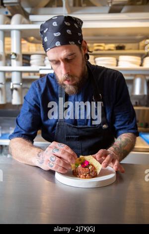 Tätowierte fokussierte männliche Koch in Schürze setzen Cracker in frischem Rindfleisch Tartare am Tisch, während der Arbeit in der Küche des Restaurants Stockfoto