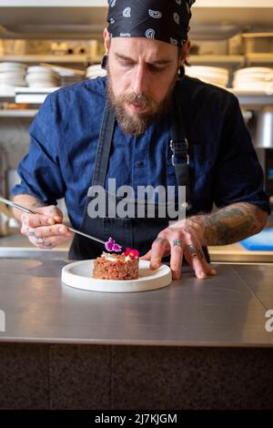 Gebärtete Chefköchin auf der Schürze, die frischen, leckeren Rindertartar auf dem Teller in der Küche des Restaurants verschönert Stockfoto