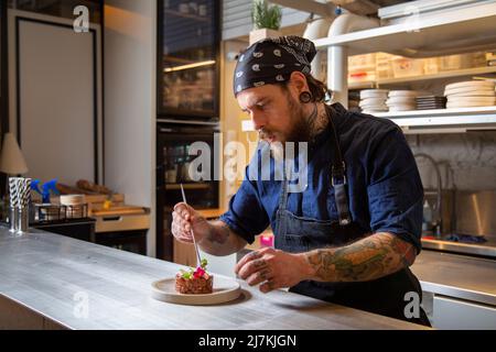 Gebärtete Chefköchin auf der Schürze, die frischen, leckeren Rindertartar auf dem Teller in der Küche des Restaurants verschönert Stockfoto
