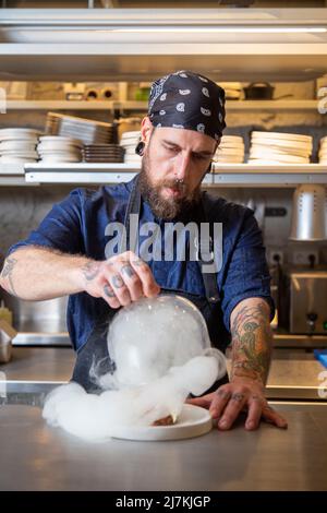 Seriös fokussierter bärtiger Chefkoch, der die Kamera anschaut, während er im Restaurant mit Rauchkanone und Glaskuppel frischen Rindertartar serviert Stockfoto