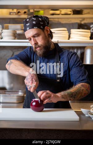 Fokussierter tätowierte Chefkoch in Schürze, der rote Zwiebeln mit Messer hackt, während er in einer hellen Küche kocht Stockfoto