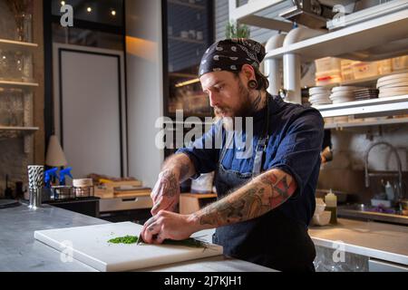 Fokussierter tätowierte Chefkoch schneidet frische grüne Zwiebeln mit scharfem Messer auf dem Schneidebrett, während er in der hellen Küche kocht Stockfoto