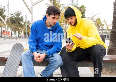 Zwei Freunde Skater mit bunten Outfits per Telefon, während sie auf einer Bank auf der Straße sitzen Stockfoto
