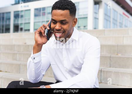 Positiver afroamerikanischer Mann mit schwarzem Haar in formeller Kleidung, der auf einer Steintreppe in der Stadt telefoniert Stockfoto