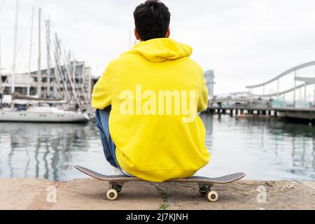 Rückansicht eines anonymen Mannes mit gelbem Hoodie, der auf Skaten vor einem Hafen sitzt Stockfoto