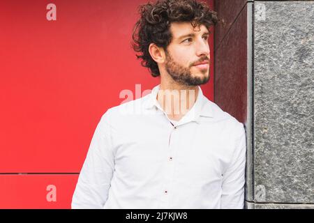 Nachdenklicher bärtiger männlicher Unternehmer mit schwarzen lockigen Haaren, der in der Nähe der roten Gebäudewand auf der Straße der Stadt steht Stockfoto